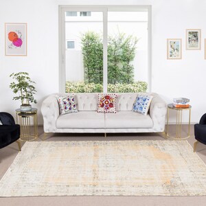 May include: A living room interior featuring a white sofa, two black armchairs, and a patterned rug. The sofa has three colourful throw pillows. The armchairs have one throw pillow each. There are two gold side tables with glass tops. The room has a large window with a view of green trees. There are framed pictures on the wall.