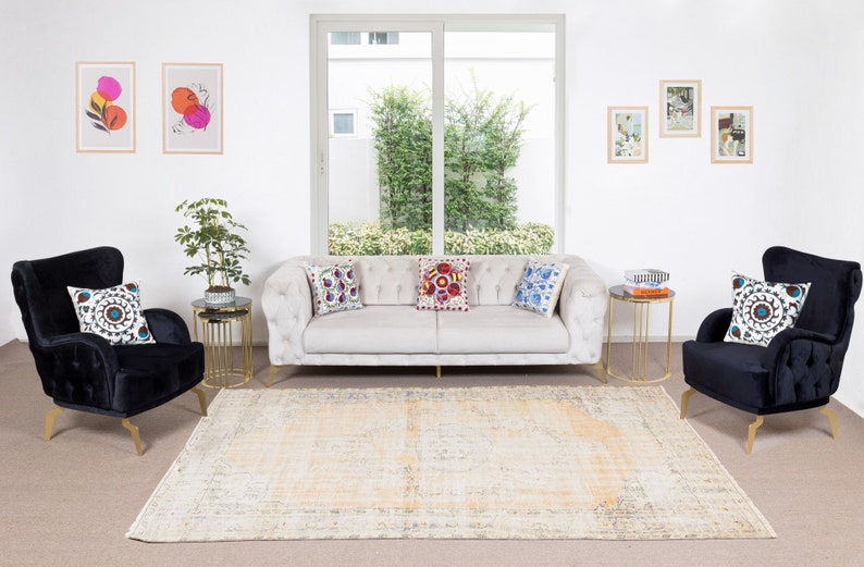 May include: A living room interior featuring a white sofa, two black armchairs, and a patterned rug. The sofa has three colourful throw pillows. The armchairs have one throw pillow each. There are two gold side tables with glass tops. The room has a large window with a view of green trees. There are framed pictures on the wall.