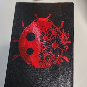 Puede incluir: Diario negro con un diseño de mariquita roja. La mariquita está dividida, con un lado que presenta un patrón floral. Un dije de mariquita roja y plateada cuelga de un marcador de cinta negra.