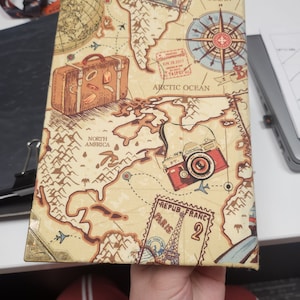 Puede incluir: Un diario de viaje con un diseño de mapa vintage. La portada presenta una rosa de los vientos, una maleta y una ilustración de cámara. El diario incluye el texto "ATLANTIC OCEAN", "ARCTIC OCEAN", "NORTH AMERICA", "REPUB FRANC PARIS 2" y un sello de fecha.