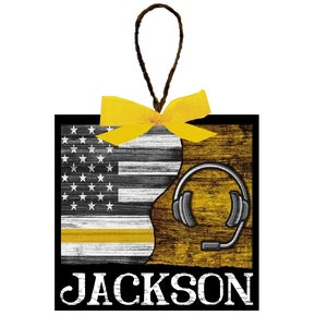 May include: A wooden ornament with a yellow ribbon, a black and white American flag, a yellow thin line, and a headset. The ornament has the name "JACKSON" printed on the bottom.