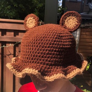 May include: A brown crocheted bucket hat with brown bear ears. The hat has a brown crocheted brim with a tan crocheted edge.