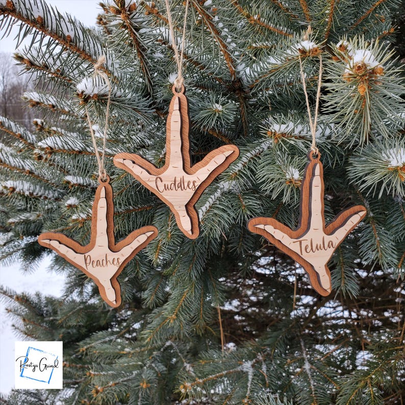 May include: Three wooden ornaments shaped like a chicken foot with the names "Cuddles", "Peaches", and "Telula" engraved on them. The ornaments are hanging on a green Christmas tree.