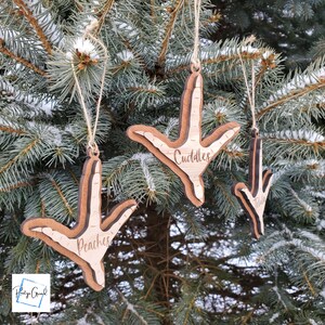 May include: Three wooden ornaments shaped like a chicken foot with the names "Cuddles", "Peaches", and "Tilda" engraved on them. The ornaments are hanging on a pine tree branch.