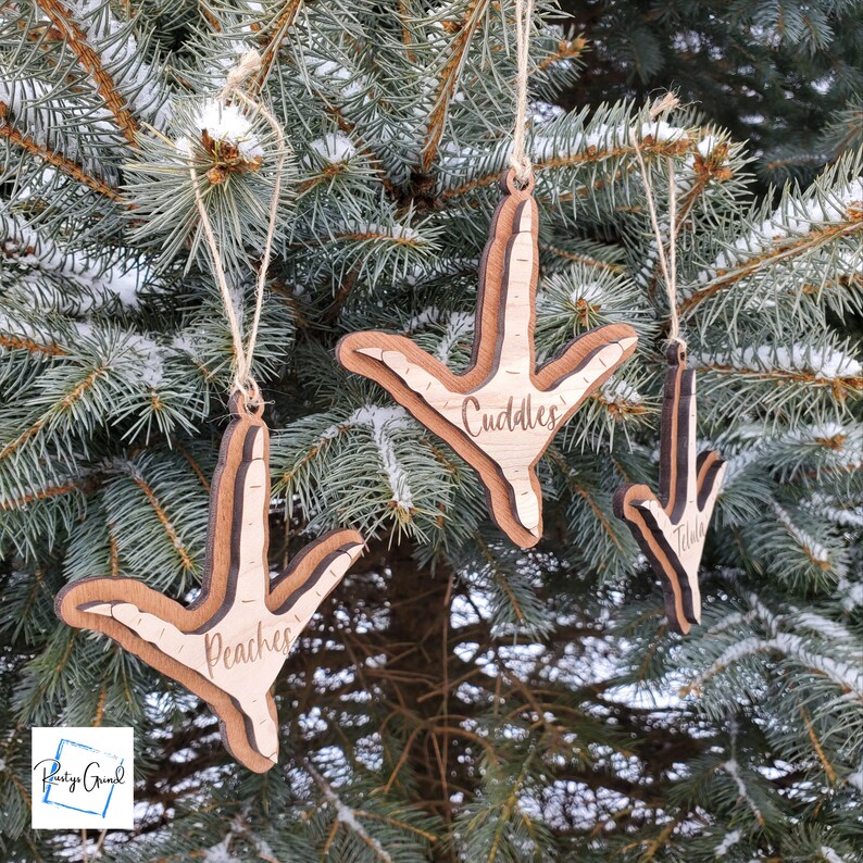 May include: Three wooden ornaments shaped like a chicken foot with the names "Cuddles", "Peaches", and "Tilda" engraved on them. The ornaments are hanging on a pine tree branch.
