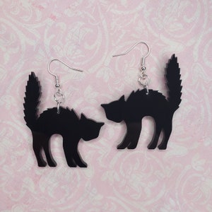 May include: A pair of black cat silhouette earrings. The earrings are made of acrylic and have silver hooks.