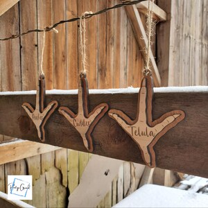 May include: Three wooden chicken foot ornaments with the names Peaches, Cuddles, and Telula engraved on them. The ornaments are hanging from a wooden beam against a snowy background.