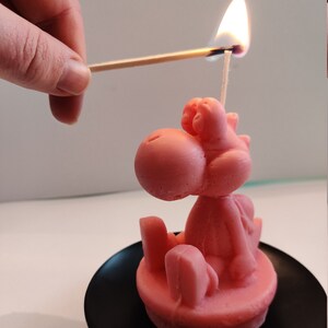 Pode incluir: Uma vela rosa em forma de personagem de desenho animado está sendo acesa com um fósforo. A vela está em um prato preto. A vela é de cor rosa sólida e tem um design detalhado.