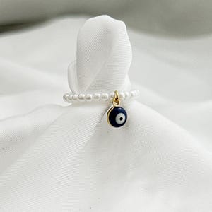 Könnte beinhalten: Ein perlenbesetzter Ring mit einem goldenen Anhänger mit einem blau-weißen bösen Blick-Design. Der Ring wird auf einem weißen Stoffhintergrund präsentiert. Der Ring ist ein Schmuckstück.