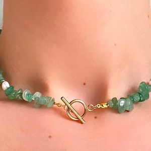 Op de afbeelding: Een groene edelsteenketting met een goudkleurige sluiting. De ketting heeft kleine, onregelmatig gevormde groene stenen en witte kralen. De sluiting is een eenvoudige tuimelsluiting. De ketting wordt om de nek gedragen.