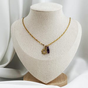Könnte beinhalten: Eine goldfarbene Halskette mit zwei kleinen Anhängern: einem kreisförmigen Sonnenmotiv mit strahlenden Strahlen und einem einzelnen, länglichen, tiefvioletten Kristall. Die feine Metallkette hat eine texturierte, reflektierende Oberfläche.