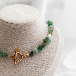 Op de afbeelding: Een groene edelsteenketting met een goudkleurige sluiting. De ketting is gemaakt van onregelmatig gevormde groene stenen en een paar witte stenen. De sluiting is een goudkleurige staaf- en ringsluiting. De ketting wordt getoond op een witte mannequin.