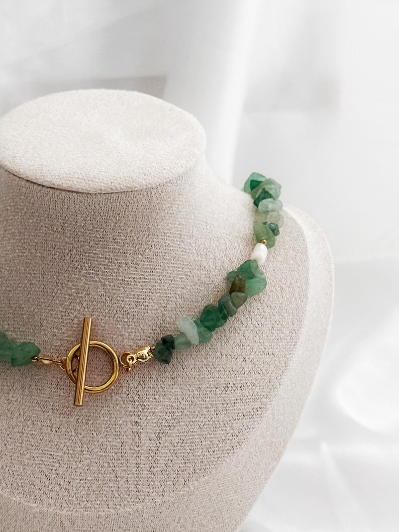 Op de afbeelding: Een groene edelsteenketting met een goudkleurige sluiting. De ketting is gemaakt van onregelmatig gevormde groene stenen en een paar witte stenen. De sluiting is een goudkleurige staaf- en ringsluiting. De ketting wordt getoond op een witte mannequin.