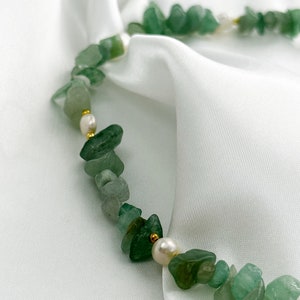 Op de afbeelding: Een ketting met onregelmatige groene stenen kralen, witte parels en kleine gouden kralen. De ketting wordt gepresenteerd op witte, zijdeachtige stof. De lengte is ongeveer 40 cm.