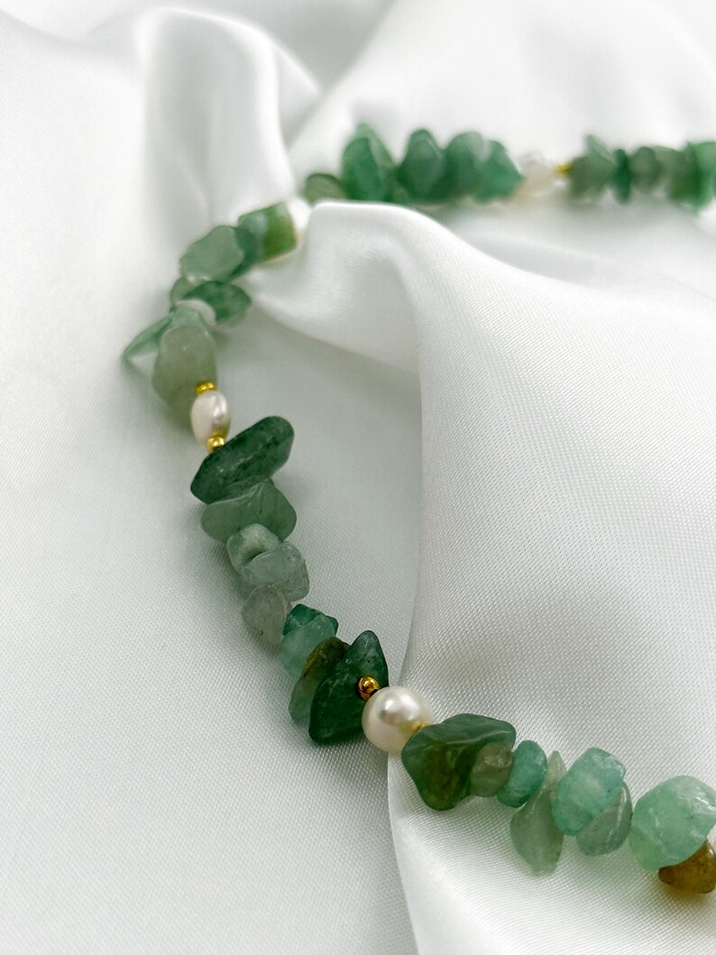 Op de afbeelding: Een ketting met onregelmatige groene stenen kralen, witte parels en kleine gouden kralen. De ketting wordt gepresenteerd op witte, zijdeachtige stof. De lengte is ongeveer 40 cm.