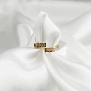 Könnte beinhalten: Zwei goldfarbene Ringe auf weißem Satin. Ein Ring hat eine Reihe kleiner, klarer Steine, der andere ein hellrosa Emailleband. Die Ringe sind offen, mit einem modernen Design. Die Ringe werden auf einem weißen Satintuch präsentiert.