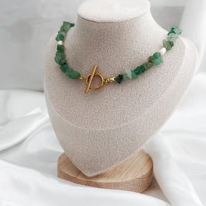 Op de afbeelding: Een groene stenen ketting met een goudkleurige sluiting wordt getoond op een mannequin. De ketting heeft onregelmatige groene stenen en kleine witte kralen. De sluiting is een eenvoudig staaf- en ringontwerp. De ketting wordt gepresenteerd op een beige met stof bedekte buste.
