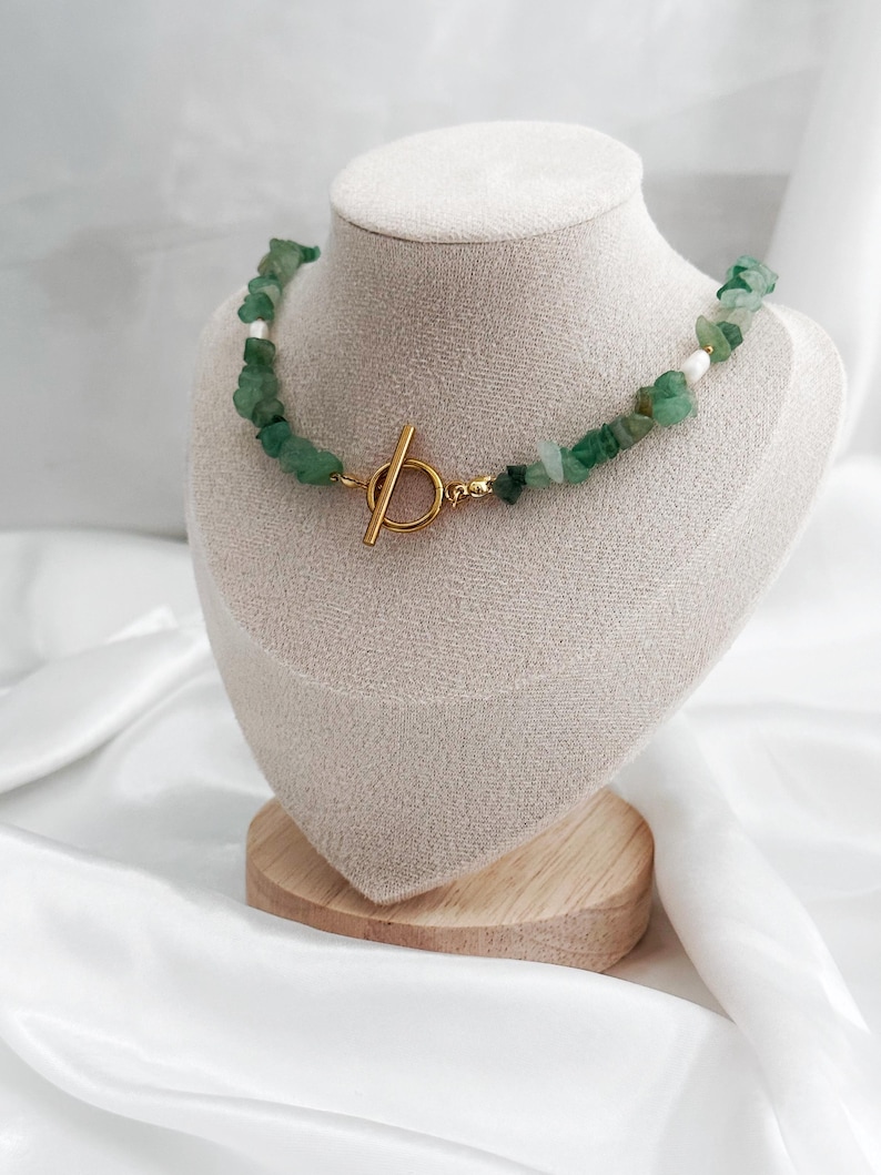 Op de afbeelding: Een groene stenen ketting met een goudkleurige sluiting wordt getoond op een mannequin. De ketting heeft onregelmatige groene stenen en kleine witte kralen. De sluiting is een eenvoudig staaf- en ringontwerp. De ketting wordt gepresenteerd op een beige met stof bedekte buste.