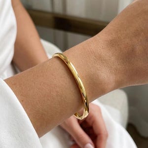 Könnte beinhalten: Ein polierter goldfarbener Armreif mit einer glatten, abgerundeten Oberfläche und einem durchgehenden kreisförmigen Design. Das Metall hat ein reflektierendes, hochglänzendes Finish, das das Licht einfängt und eine schlichte Ästhetik bietet.
