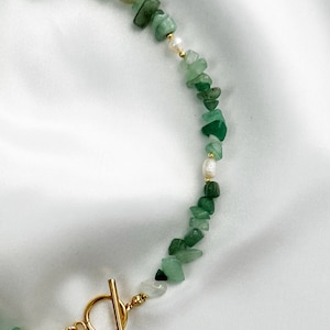 Op de afbeelding: Een ketting met groene aventurijn chip kralen en kleine witte parels. De ketting heeft een goudkleurige toggle sluiting. De kralen zijn geregen op een delicate ketting, wat een natuurlijke en elegante uitstraling cre&euml;ert.