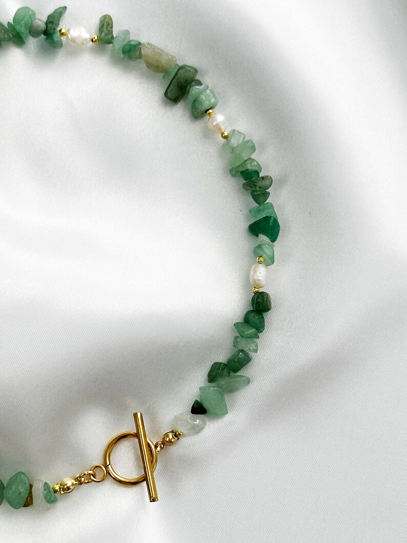 Op de afbeelding: Een ketting met groene aventurijn chip kralen en kleine witte parels. De ketting heeft een goudkleurige toggle sluiting. De kralen zijn geregen op een delicate ketting, wat een natuurlijke en elegante uitstraling cre&euml;ert.
