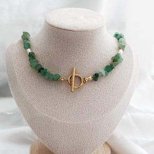Op de afbeelding: Een groene edelsteenketting met een goudkleurige sluiting. De ketting wordt op een mannequin getoond. De edelstenen zijn een mix van groen en wit. De sluiting is een T-bar stijl.