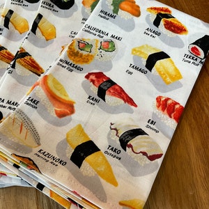 May include: A white fabric napkin with a colourful illustration of different types of sushi. The napkin features the names of each type of sushi in English, including "Ikura", "Hirame", "California Maki", "Anago", "Tamago", "Tekka Maki", "Sake", "W/Masago", "Kani", "Ebi", "Kazunoko", and "Tako".