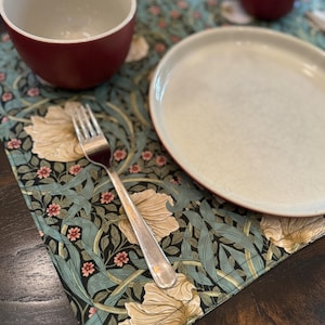 May include: A place setting with a dark green and cream floral patterned placemat, a white plate, a silver fork, and a burgundy bowl.