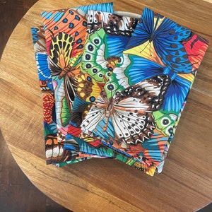 May include: A stack of colourful fabric napkins with a butterfly pattern. The napkins are folded and arranged on a wooden surface.