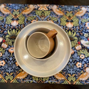 May include: A set of three beige ceramic dishes, including a small cup and two larger plates, on a blue and green floral patterned placemat.