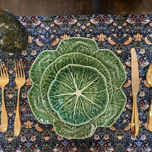 May include: A table setting featuring a green cabbage-shaped plate set, gold flatware, and a patterned placemat with birds and floral designs. A clear, textured glass is also present. The arrangement is set on a dark wooden surface.