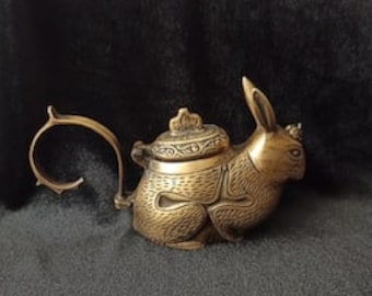 May include: A bronze-colored teapot in the shape of a rabbit. The teapot has a curved handle and a lid with a decorative finial. The rabbit is in a seated position with its ears perked up. The teapot is on a dark gray surface.