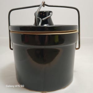May include: A black ceramic crock with a lid and a metal handle. The crock has a glossy finish and a cylindrical shape. The lid has a small, rounded knob. The metal handle is attached to the sides of the crock.