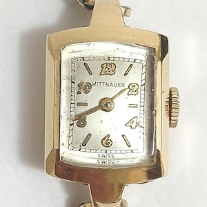May include: A vintage gold-tone Wittnauer wristwatch featuring a rectangular case with a silver dial, gold-colored hands, and gold-tone Arabic numerals at the 12, 2, 4, 6, 8, and 10 o'clock positions. The word SWISS is printed at the bottom of the dial.