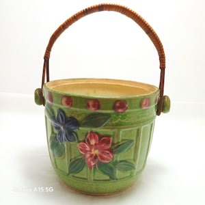 May include: A green ceramic basket with a woven handle. The basket features embossed floral designs in red, blue, and green. The rim is decorated with red dots. A charming decorative piece for home decor.