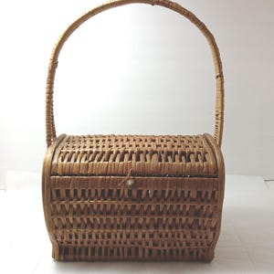 May include: A woven wicker handbag with a curved handle. The rectangular body has rounded sides and a small clasp. The basket is a light brown color and appears to be vintage.