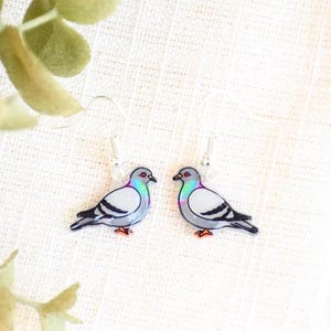 May include: A pair of silver-toned earrings featuring a pigeon design. Each earring showcases a gray bird with black and white details, a red beak and feet, and a colorful iridescent chest. The earrings hang from silver-colored hooks.