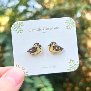 May include: A pair of handmade duck earrings on a white card with the text "Camille Christine Art" and "Handmade Earrings". The earrings are yellow and brown, with a duck design. The background is blurred green.