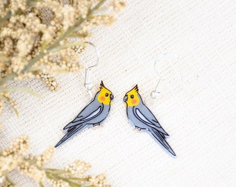 May include: A pair of silver dangle earrings featuring a gray and yellow cockatiel design.