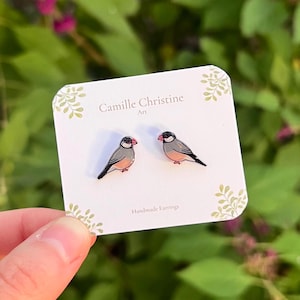 May include: A pair of stud earrings featuring illustrations of Java sparrows with grey, black, and pink plumage. The earrings are attached to a white display card printed with the text Camille Christine Art and Handmade Earrings.