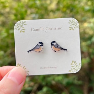 May include: A pair of stud earrings shaped like small birds with black heads, white cheeks, and tan bodies. The earrings are attached to a white display card featuring the text Camille Christine Art and Handmade Earrings.