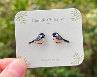 May include: A pair of stud earrings shaped like small birds with black heads, white cheeks, and tan bodies. The earrings are attached to a white display card featuring the text Camille Christine Art and Handmade Earrings.