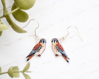 May include: A pair of earrings featuring a detailed illustration of a kestrel bird with orange, brown, and blue colors. The earrings are made of clear acrylic and have silver hooks.