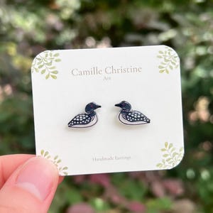 May include: A pair of handmade earrings featuring a loon design. The earrings are black and white with a detailed pattern, presented on a white card with the text "Camille Christine Art" and "Handmade Earrings".