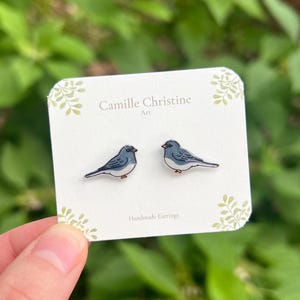 Può includere: Un paio di orecchini a lobo con illustrazioni di piccoli uccelli blu e grigi con becchi arancioni. Gli orecchini sono fissati su un cartoncino rettangolare bianco con stampato il testo Camille Christine Art e Handmade Earrings.