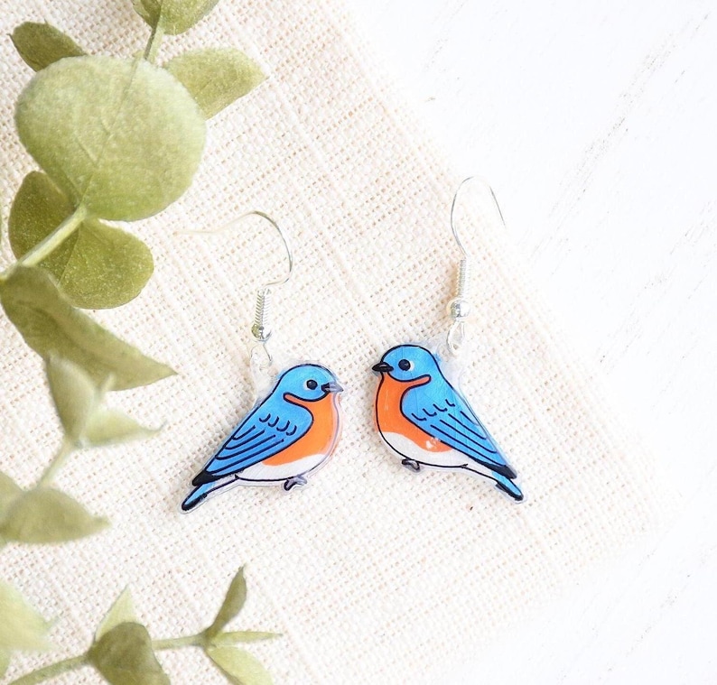 May include: A pair of bluebird earrings with silver hooks. The bluebirds are made of clear acrylic with a blue, orange, and white painted design.