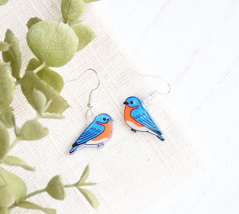 May include: A pair of bluebird earrings with silver hooks. The bluebirds are painted with blue, orange, and black details.