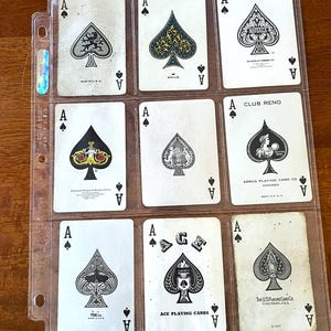 May include: A collection of nine vintage ace of spades playing cards displayed in a clear plastic protective sheet. Each card features a unique black and white spade design, including various brand logos and intricate illustrations of figures and patterns.