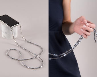 May include: A silver phone case with a matching beaded lanyard. The lanyard is made of small, square-shaped beads. The phone case is clear and attached to the lanyard with silver hardware. The phone case is resting on a white marble stand.