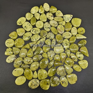 May include: A collection of green and black speckled gemstone cabochons. The cabochons are in various shapes and sizes, with some being oval, teardrop, and round. The stones are a mix of light and dark green, with black speckles throughout.